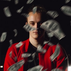 A person wearing a red jacket is surrounded by fluttering dollar bills against a dark background. The lighting casts deep shadows, highlighting the person's face while the bills are in motion, creating a dramatic and dynamic scene.