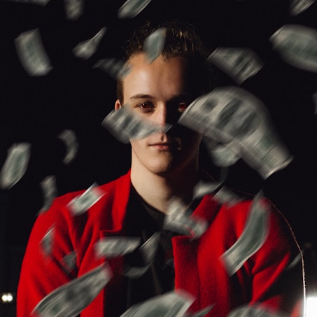 A person wearing a red jacket is surrounded by fluttering dollar bills against a dark background. The lighting casts deep shadows, highlighting the person's face while the bills are in motion, creating a dramatic and dynamic scene.