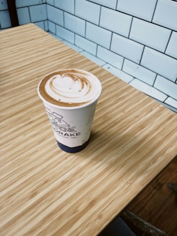 A cup of coffee with latte art sits on a light brown wooden table. Behind the table is a wall made of white subway tiles. The cup has a sleeve with a design, and the coffee has a swirl pattern on top.