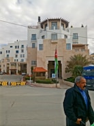 A multi-story building with modern architecture and white walls. There's a Jordanian flag prominently displayed near the entrance. The building is surrounded by minimal landscaping with some palm trees. A man is walking in the foreground wearing a blue shirt and dark jacket. A blue Mercedes truck is visible, parked near the entrance.