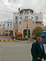 A multi-story building with modern architecture and white walls. There's a Jordanian flag prominently displayed near the entrance. The building is surrounded by minimal landscaping with some palm trees. A man is walking in the foreground wearing a blue shirt and dark jacket. A blue Mercedes truck is visible, parked near the entrance.