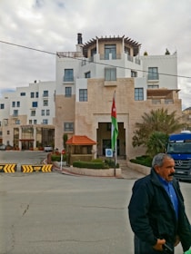 A multi-story building with modern architecture and white walls. There's a Jordanian flag prominently displayed near the entrance. The building is surrounded by minimal landscaping with some palm trees. A man is walking in the foreground wearing a blue shirt and dark jacket. A blue Mercedes truck is visible, parked near the entrance.