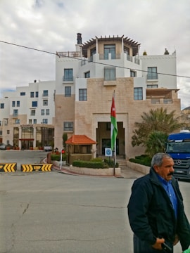 A multi-story building with modern architecture and white walls. There's a Jordanian flag prominently displayed near the entrance. The building is surrounded by minimal landscaping with some palm trees. A man is walking in the foreground wearing a blue shirt and dark jacket. A blue Mercedes truck is visible, parked near the entrance.