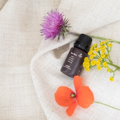 A small, brown bottle of tea tree essential oil is placed on a textured cream-colored fabric, surrounded by vibrant flowers. A purple thistle, a cluster of small yellow flowers, and a bright red poppy are artfully arranged around the bottle, creating a visually appealing composition.