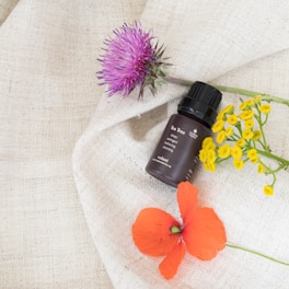 A small, brown bottle of tea tree essential oil is placed on a textured cream-colored fabric, surrounded by vibrant flowers. A purple thistle, a cluster of small yellow flowers, and a bright red poppy are artfully arranged around the bottle, creating a visually appealing composition.