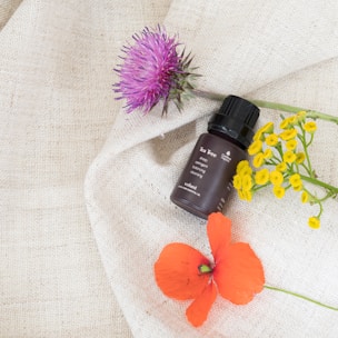 A small, brown bottle of tea tree essential oil is placed on a textured cream-colored fabric, surrounded by vibrant flowers. A purple thistle, a cluster of small yellow flowers, and a bright red poppy are artfully arranged around the bottle, creating a visually appealing composition.