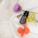 A small, brown bottle of tea tree essential oil is placed on a textured cream-colored fabric, surrounded by vibrant flowers. A purple thistle, a cluster of small yellow flowers, and a bright red poppy are artfully arranged around the bottle, creating a visually appealing composition.
