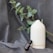 A white, sleek essential oil diffuser emits a gentle wisp of vapor, placed alongside a small dark bottle labeled ‘Eucalyptus’. They both rest on a soft gray fabric, accompanied by a branch of eucalyptus leaves that adds a natural touch.