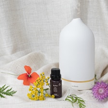 A white ceramic diffuser with a minimalist design is placed on a textured fabric backdrop. In front of the diffuser, there is a small dark bottle labeled 'Tea Tree' and 'volant' along with botanical descriptions. Various colorful flowers and green leaves are artfully arranged around the bottle and diffuser, including an orange flower, yellow blossoms, and a purple thistle-like flower.