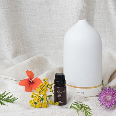 A white ceramic diffuser with a minimalist design is placed on a textured fabric backdrop. In front of the diffuser, there is a small dark bottle labeled 'Tea Tree' and 'volant' along with botanical descriptions. Various colorful flowers and green leaves are artfully arranged around the bottle and diffuser, including an orange flower, yellow blossoms, and a purple thistle-like flower.