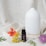 A white ceramic diffuser with a minimalist design is placed on a textured fabric backdrop. In front of the diffuser, there is a small dark bottle labeled 'Tea Tree' and 'volant' along with botanical descriptions. Various colorful flowers and green leaves are artfully arranged around the bottle and diffuser, including an orange flower, yellow blossoms, and a purple thistle-like flower.