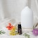 A white ceramic diffuser with a minimalist design is placed on a textured fabric backdrop. In front of the diffuser, there is a small dark bottle labeled 'Tea Tree' and 'volant' along with botanical descriptions. Various colorful flowers and green leaves are artfully arranged around the bottle and diffuser, including an orange flower, yellow blossoms, and a purple thistle-like flower.
