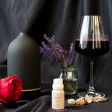 A matte black diffuser emits a subtle stream of smoke next to a bottle labeled 'Date Night.' In front, a single red rose lies on a dark satin surface. A small vase with lavender sprigs and a glass of red wine complement the elegant setup. Crystals or stones are scattered around, adding a natural element.