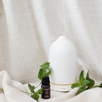 A white ceramic essential oil diffuser sits on a textured beige cloth surface, accompanied by a small dark glass bottle labeled 'Spearmint'. Fresh green mint leaves are scattered around, enhancing the natural and soothing aesthetic.