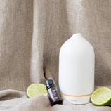 A white ceramic diffuser is placed on a textured beige fabric background, accompanied by a small dark glass bottle labeled 'Essential Oil'. Two lime halves are positioned around the objects.