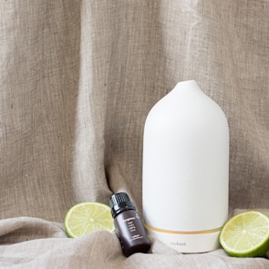 A white ceramic diffuser is placed on a textured beige fabric background, accompanied by a small dark glass bottle labeled 'Essential Oil'. Two lime halves are positioned around the objects.