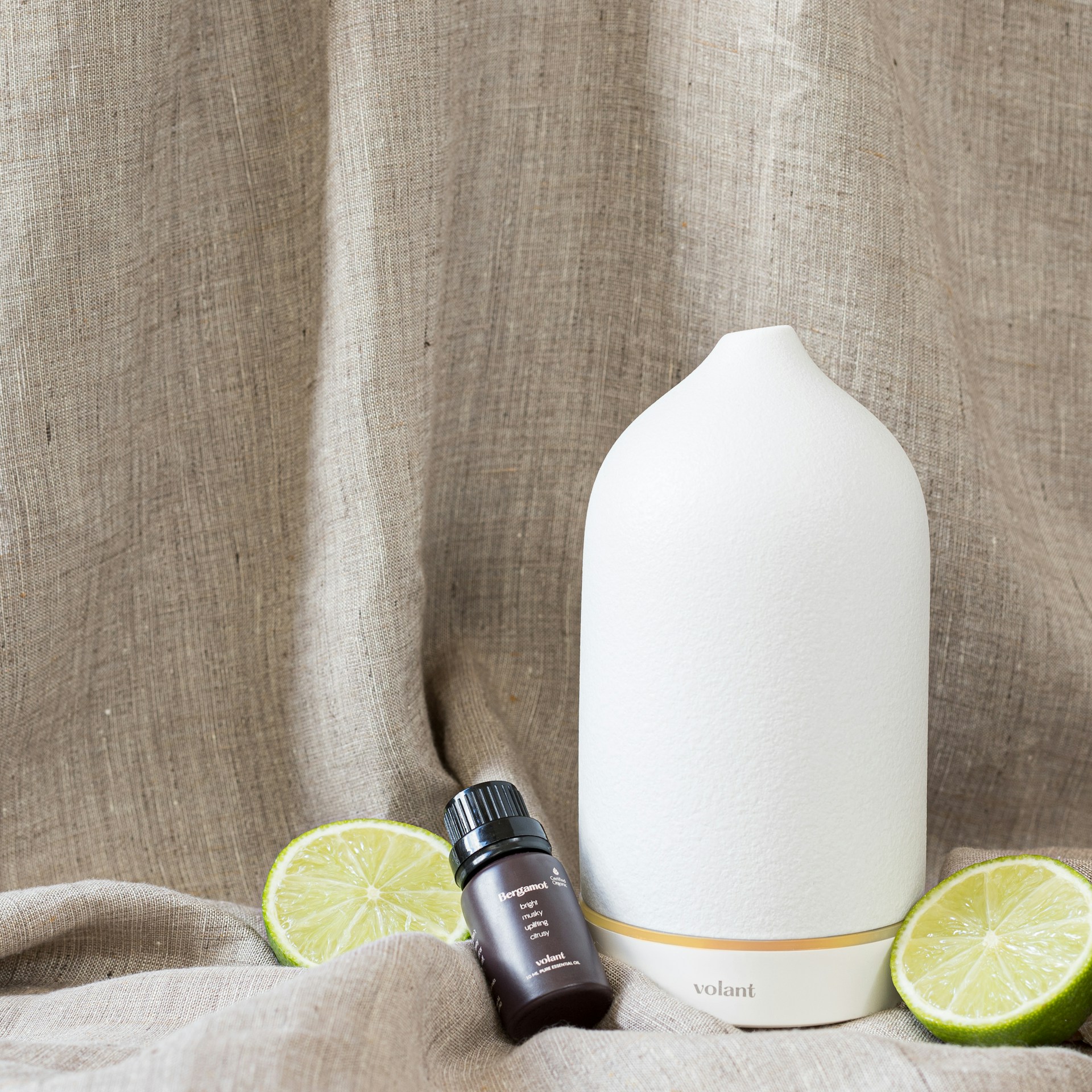 A white ceramic diffuser is placed on a textured beige fabric background, accompanied by a small dark glass bottle labeled 'Essential Oil'. Two lime halves are positioned around the objects.
