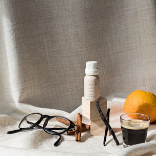 A still life composition featuring a small bottle of essential oil with a light label, placed on wooden blocks. Nearby, there are a pair of black glasses, a whole orange, a few cinnamon sticks, and a vanilla pod. A small glass of espresso is set to the right. The background and surface are draped with a textured, light fabric, creating a warm and cozy ambiance.