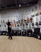 A room with electric guitars displayed on a wall, each hanging on individual hooks. Below the guitars are several amplifiers in a line. A person stands in front of the wall, gesturing as if inspecting or choosing a guitar. The floor is made of wood, and the room is lit with overhead lighting.