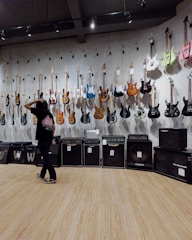 A room with electric guitars displayed on a wall, each hanging on individual hooks. Below the guitars are several amplifiers in a line. A person stands in front of the wall, gesturing as if inspecting or choosing a guitar. The floor is made of wood, and the room is lit with overhead lighting.