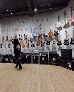 A room with electric guitars displayed on a wall, each hanging on individual hooks. Below the guitars are several amplifiers in a line. A person stands in front of the wall, gesturing as if inspecting or choosing a guitar. The floor is made of wood, and the room is lit with overhead lighting.