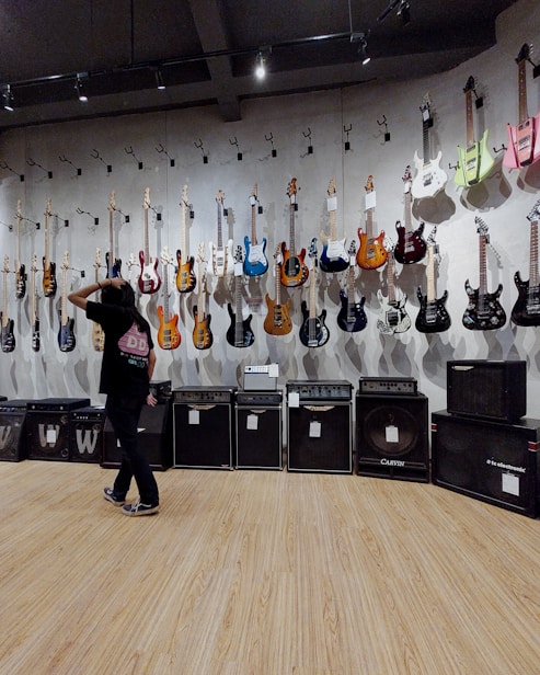 A room with electric guitars displayed on a wall, each hanging on individual hooks. Below the guitars are several amplifiers in a line. A person stands in front of the wall, gesturing as if inspecting or choosing a guitar. The floor is made of wood, and the room is lit with overhead lighting.