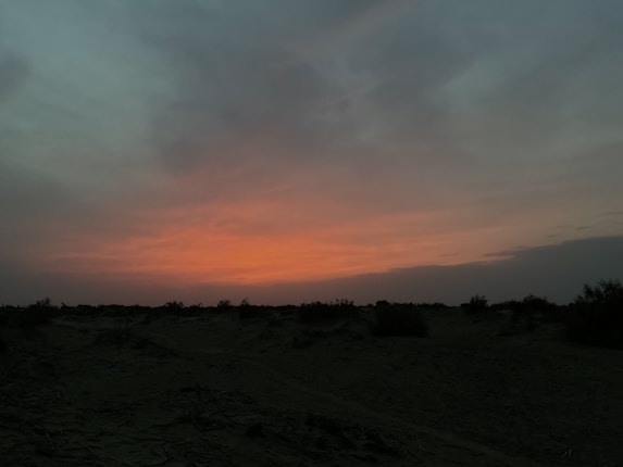 A serene landscape featuring a vivid sunset over an expansive desert. The sky is filled with warm orange and pink hues that fade into darker blues and grays. The foreground is shadowed with silhouettes of sparse desert vegetation.