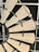 A close-up view of a windmill style ceiling fan with wooden blades. The fan blades are attached to a central hub with black brackets. The background shows the metal framework of a ceiling grid.
