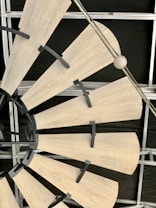 A close-up view of a windmill style ceiling fan with wooden blades. The fan blades are attached to a central hub with black brackets. The background shows the metal framework of a ceiling grid.