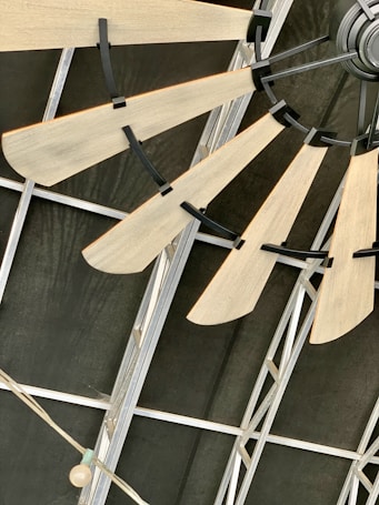 Wooden fan blades attached to a central hub mounted on an industrial-style metal ceiling. The structure consists of metal beams and a string of lights with a single exposed bulb.