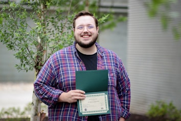 A person wearing glasses and a plaid shirt is smiling while holding an open folder with a certificate inside. The background features a small tree and blurred outdoor elements.