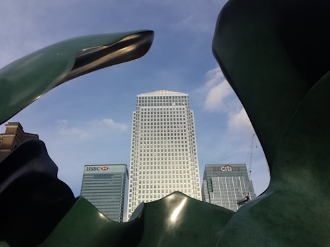A modern cityscape featuring prominent skyscrapers with expansive glass facades. The foreground includes a large, abstract green sculpture framing the buildings, which include HSBC and Citi towers. The sky is clear with patches of blue and a few clouds.