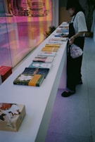 A person wearing a mask and dark clothing stands beside a long white table displaying various books with colorful covers. The background features a vibrant, reflective surface with a distorted image of a truck and street scene, adding a dynamic touch to the setting.