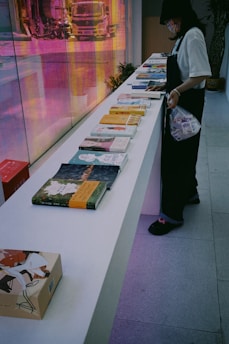A person wearing a mask and dark clothing stands beside a long white table displaying various books with colorful covers. The background features a vibrant, reflective surface with a distorted image of a truck and street scene, adding a dynamic touch to the setting.