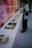 A person wearing a mask and dark clothing stands beside a long white table displaying various books with colorful covers. The background features a vibrant, reflective surface with a distorted image of a truck and street scene, adding a dynamic touch to the setting.