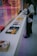 A person wearing a mask and dark clothing stands beside a long white table displaying various books with colorful covers. The background features a vibrant, reflective surface with a distorted image of a truck and street scene, adding a dynamic touch to the setting.