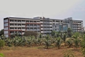 A large, multi-story building with a modern architectural design featuring a combination of red, white, and gray colors. The structure is surrounded by greenery, including palm trees in the foreground, and appears to be located in a semi-rural setting.