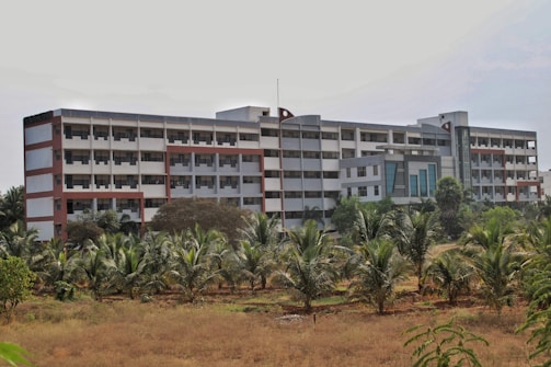 A large, multi-story building with a modern architectural design featuring a combination of red, white, and gray colors. The structure is surrounded by greenery, including palm trees in the foreground, and appears to be located in a semi-rural setting.