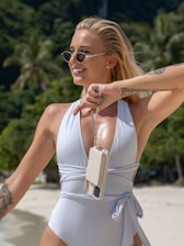 A woman wearing sunglasses and a white swimsuit stands on a sunny beach, smiling and holding a phone in a case hanging from her wrist. Her hair is blonde and tied back, and she has tattoos on her arms. The background features palm trees and sand.