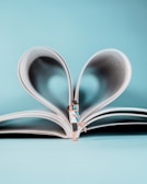 A miniature couple embraces joyfully in front of an open book, whose pages are folded into a heart shape. The background is a soft, pastel blue, enhancing the romantic and whimsical nature of the scene.