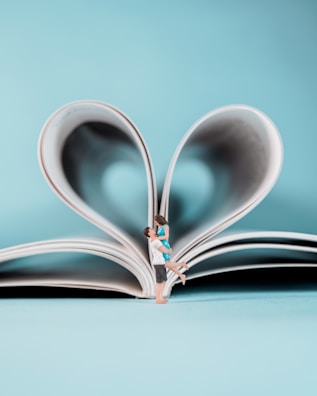 A miniature couple embraces joyfully in front of an open book, whose pages are folded into a heart shape. The background is a soft, pastel blue, enhancing the romantic and whimsical nature of the scene.