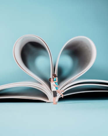 A miniature couple embraces joyfully in front of an open book, whose pages are folded into a heart shape. The background is a soft, pastel blue, enhancing the romantic and whimsical nature of the scene.