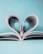A miniature couple embraces joyfully in front of an open book, whose pages are folded into a heart shape. The background is a soft, pastel blue, enhancing the romantic and whimsical nature of the scene.