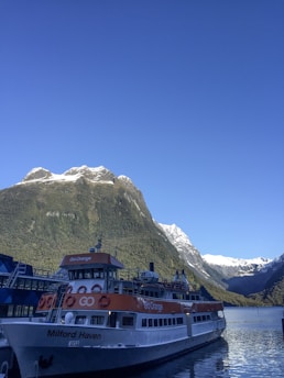 A large ferry named Milford Haven is docked at a pier with towering snow-capped mountains in the background under a clear blue sky. The ferry has a distinctive orange and white color scheme and is part of the Go Orange fleet. The mountains provide a dramatic and scenic backdrop, and the calm water reflects the serene atmosphere.