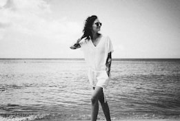 A person stands by the edge of the sea, wearing sunglasses and a light, casual outfit. The person has tattoos on their arms and appears relaxed, with one hand on their head. The ocean stretches out in the background, and the scene is captured in black and white.