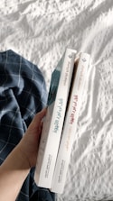 A hand holding two books with Arabic titles on the spines. The books are positioned vertically side by side, and are held over a white quilted fabric with a blue plaid cloth nearby.