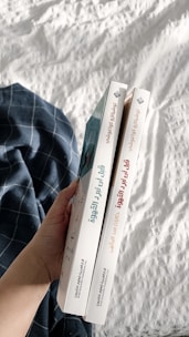 A hand holding two books with Arabic titles on the spines. The books are positioned vertically side by side, and are held over a white quilted fabric with a blue plaid cloth nearby.
