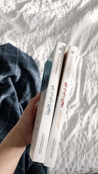 A hand holding two books with Arabic titles on the spines. The books are positioned vertically side by side, and are held over a white quilted fabric with a blue plaid cloth nearby.