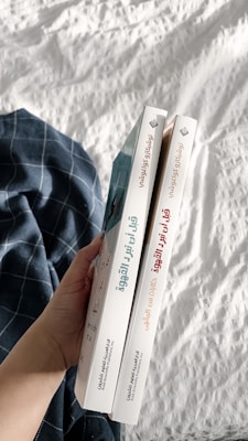 A hand holding two books with Arabic titles on the spines. The books are positioned vertically side by side, and are held over a white quilted fabric with a blue plaid cloth nearby.