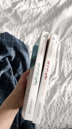 A hand holding two books with Arabic titles on the spines. The books are positioned vertically side by side, and are held over a white quilted fabric with a blue plaid cloth nearby.